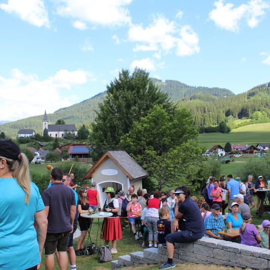 Eine Gruppe von Menschen versammelt sich im Freien, einige sitzen um Tische herum, andere stehen, mit einem kleinen Haus und einer Kirche im Hintergrund, vor einer Kulisse von Bergen und blauem Himmel.