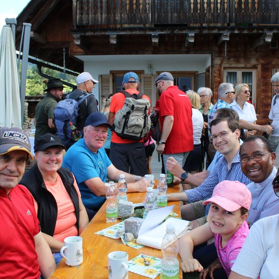 Eine Gruppe von Menschen versammelt sich um einen Tisch, einige sitzen und andere stehen, mit einem Holzhaus im Hintergrund. Sie lächeln und scheinen eine Mahlzeit oder ein Treffen zu genießen.