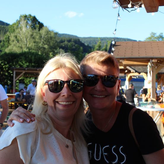 Zwei Frauen mit Sonnenbrillen lächeln für ein Foto in einer Außenumgebung mit Bergen im Hintergrund.