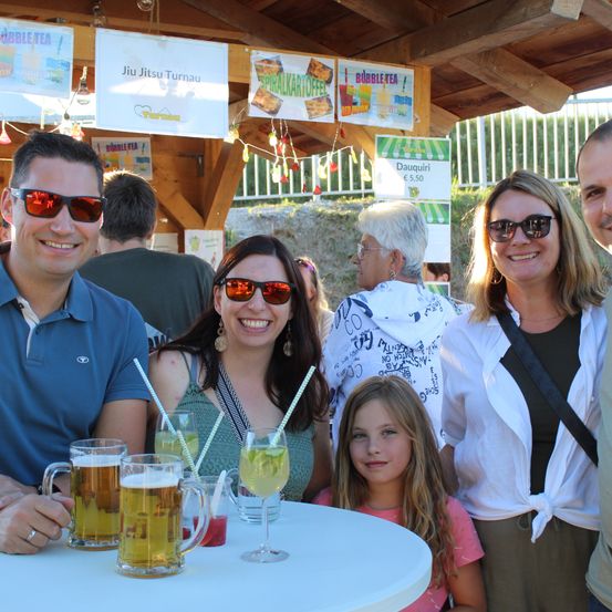 Eine vierköpfige Familie, bestehend aus zwei Erwachsenen und zwei Kindern, lächelt und posiert für ein Foto an einem Tisch mit Getränken vor einem hölzernen Pavillon mit Schildern für Bubble Tea und Jiu Jitsu.
