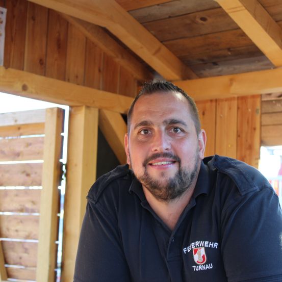 Ein Mann mit Bart trägt ein schwarzes Poloshirt mit dem Wort 'Feuerwehr' darauf und lächelt in die Kamera. Er steht in einem Holzraum mit Holzwänden und -decke.