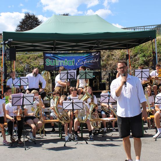 Eine Blechbläsergruppe spielt unter einem grünen Zelt, wobei ein Mann in ein Mikrofon spricht. Die Bandmitglieder sitzen auf Bänken und spielen verschiedene Blechblasinstrumente.