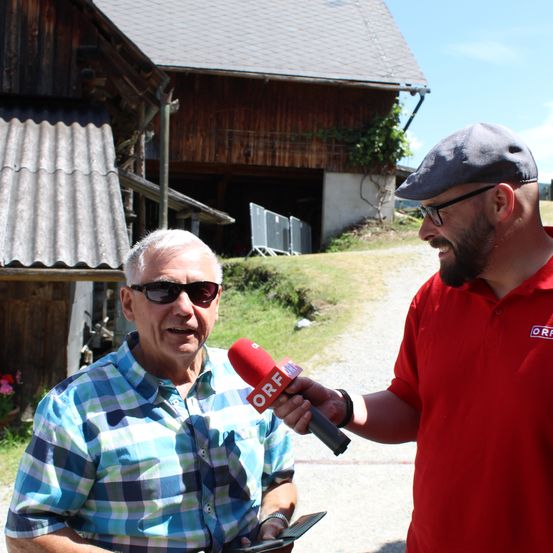 Ein Mann in einem blau karierten Hemd wird von einem Mann in einem roten Hemd und einer grauen Mütze interviewt, beide tragen Sonnenbrillen. Das Interview findet vor einem rustikalen Gebäude im Freien statt.