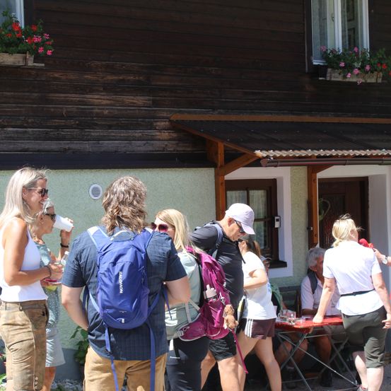 Eine Gruppe von Menschen steht vor einem Holzgebäude mit Blumenkästen, einige trinken und andere unterhalten sich. Ein Mann mit Rucksack schaut etwas nach links.