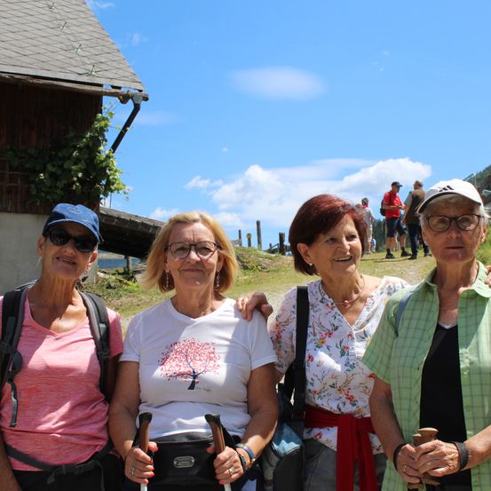 Vier Frauen in Wanderausrüstung lächeln für die Kamera, vor einem Haus mit bewölktem Himmel im Hintergrund.