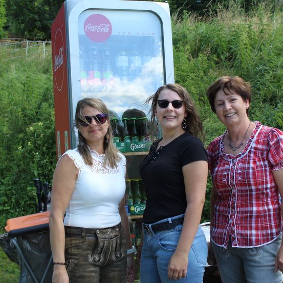 Drei Frauen stehen vor einem Coca-Cola-Automaten, lächeln und tragen Sonnenbrillen. Der Automat ist mit Flaschen von Coca-Cola und Güsser bestückt.