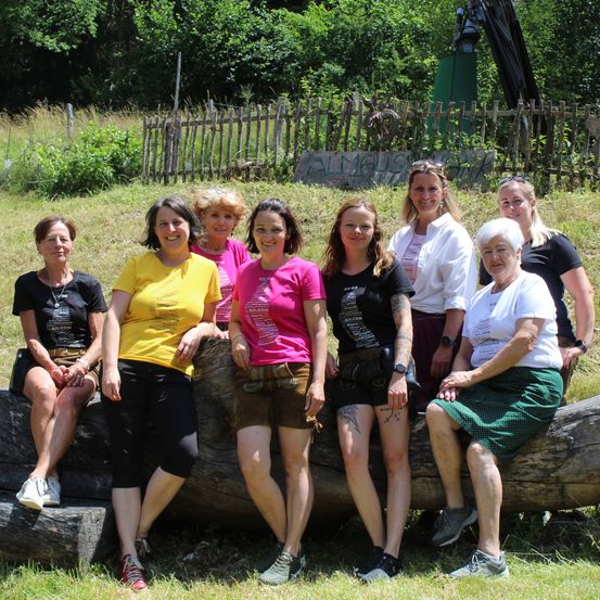 Eine Gruppe von Frauen in Freizeitkleidung sitzt auf einem Baumstamm in einem Grasfeld, lächelt und posiert für ein Foto. Hinter ihnen steht ein Holzzaun und einige Bäume.