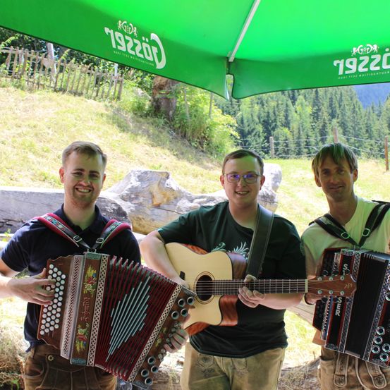 Drei Männer spielen Instrumente unter einem grünen Regenschirm. Der Mann auf der linken Seite spielt Akkordeon, der Mann in der Mitte spielt Gitarre und der Mann auf der rechten Seite spielt ebenfalls Akkordeon. Sie lächeln und scheinen für ein Foto zu posieren.
