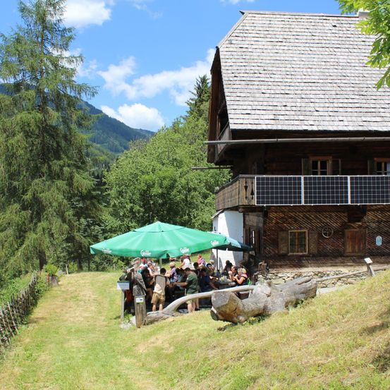 Eine Gruppe von Menschen sitzt um einen Tisch unter einem grünen Schirm in einer malerischen Outdoor-Umgebung mit einem rustikalen Haus und Bergen im Hintergrund.