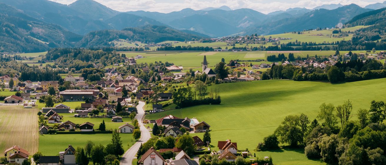 Luftaufnahme eines Dorfes in grünen Hügeln mit einer Kirche und Bergen im Hintergrund.
