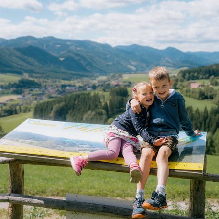 Ein Junge und ein Mädchen sitzen auf einer Holzplatte und lächeln und posieren für ein Foto. Sie sind von einer wunderschönen Berglandschaft umgeben.