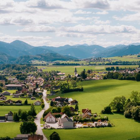 Eine Luftaufnahme eines ländlichen Dorfes, umgeben von grünen Feldern und Bergen unter einem bewölkten Himmel.