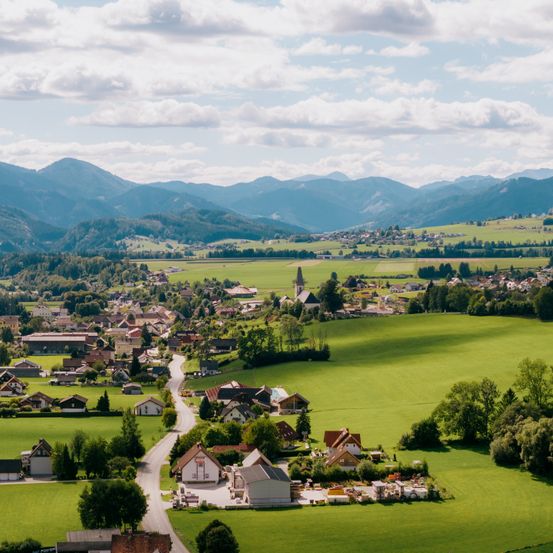 Eine Luftaufnahme eines ländlichen Dorfes, umgeben von grünen Feldern und Bergen unter einem bewölkten Himmel.