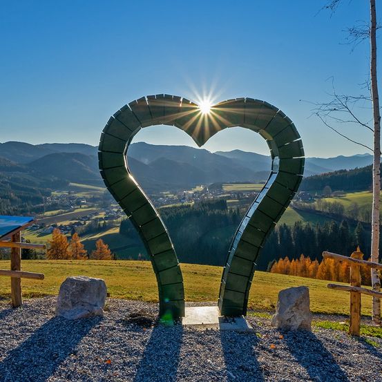 Eine herzförmige grüne Skulptur steht auf einem Hügel, mit der Sonne, die durch das Herz scheint. Berge und Bäume umgeben das Gebiet.