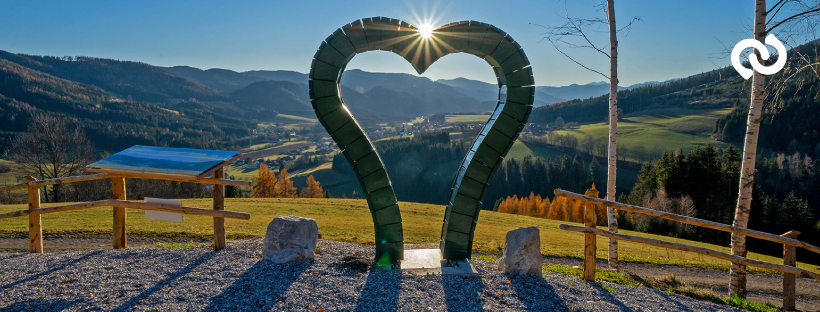 Eine große grüne herzförmige Skulptur steht auf einem Schotterbereich mit einem Holzzaun, umgeben von Bergen, grünen Hügeln und Bäumen unter einem hellen Sonnenschein.