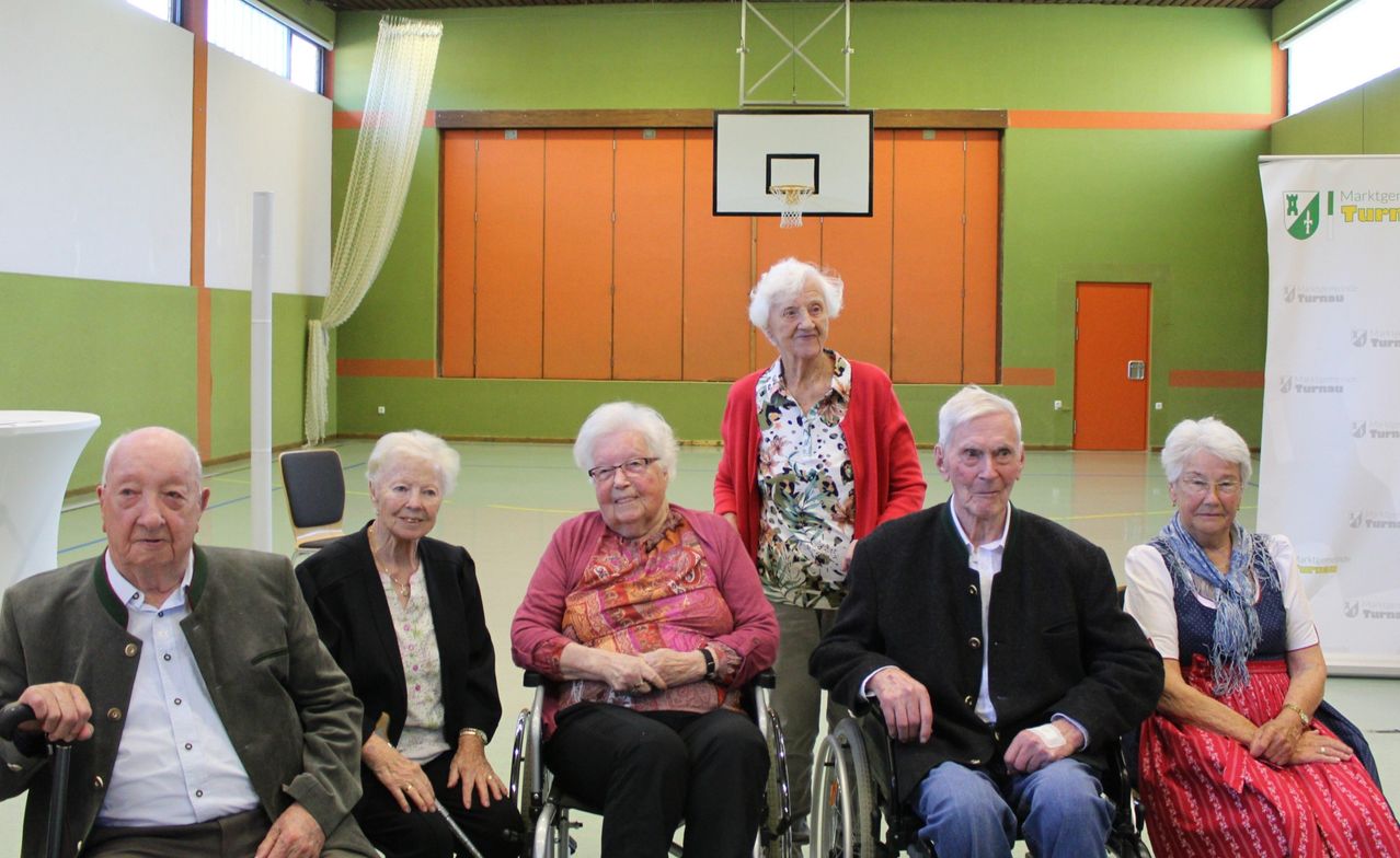 Eine Gruppe älterer Menschen in Rollstühlen posiert für ein Foto in einer Turnhalle mit einem Basketballkorb und orangefarbenen Wänden.