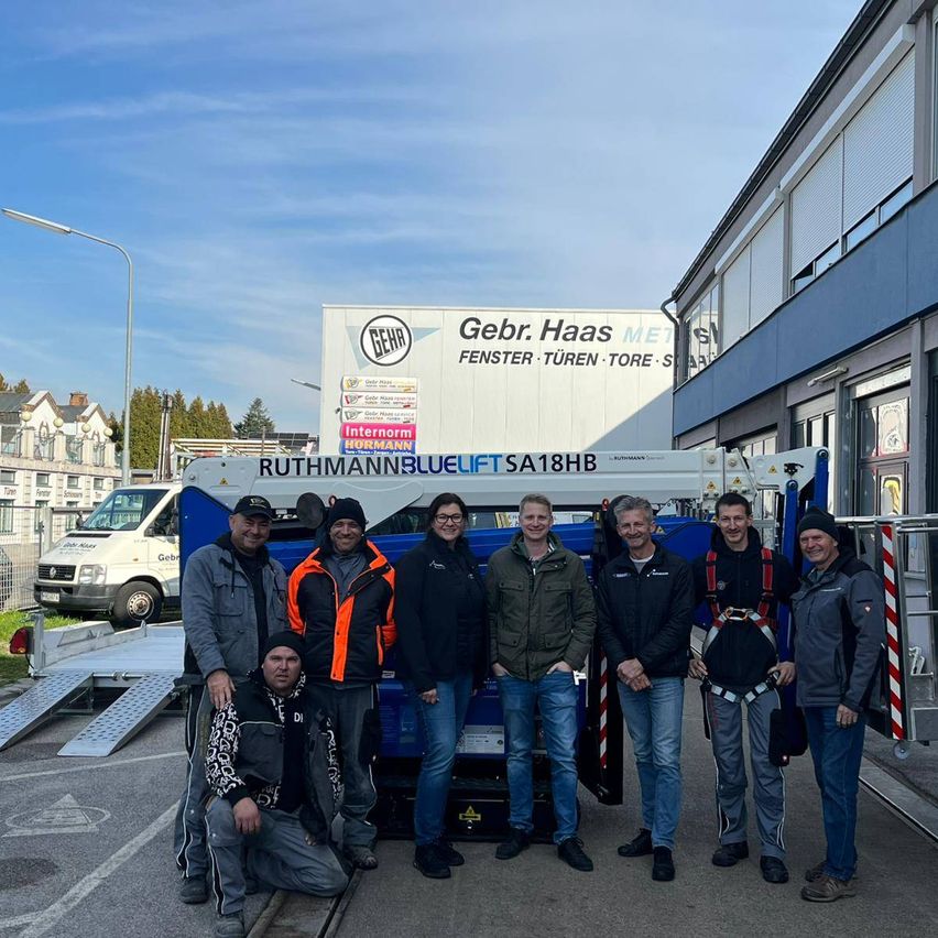 Eine Gruppe von Menschen steht vor einem blau-weißen Fahrzeug. Hinter ihnen befindet sich ein Gebäude mit einem Schild, auf dem Gebr. Haas steht. Es gibt auch andere Fahrzeuge und Straßenlaternen.