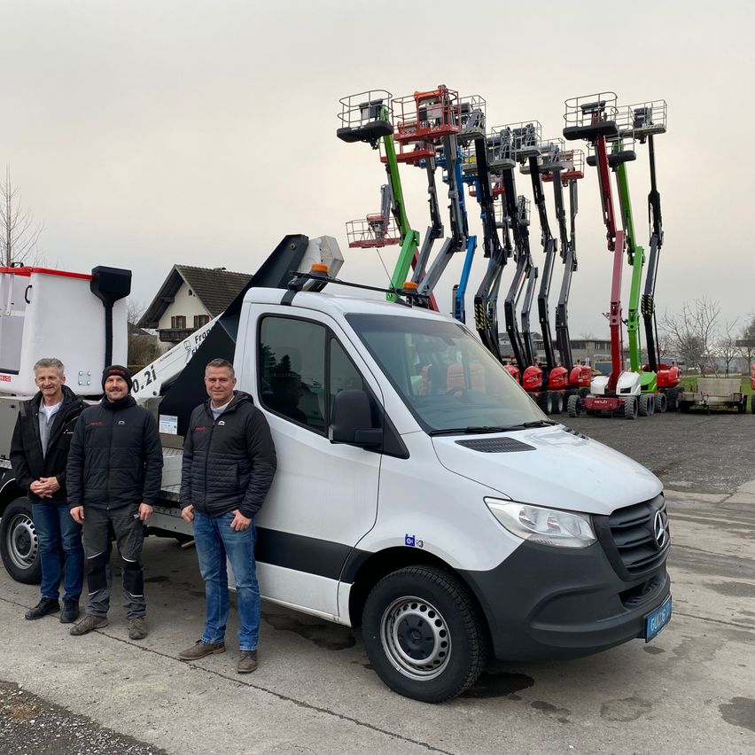 Drei Männer stehen vor einem weißen Mercedes-Lkw mit ausgefahrenem Kran, mit mehreren Kränen in verschiedenen Farben und Größen dahinter.