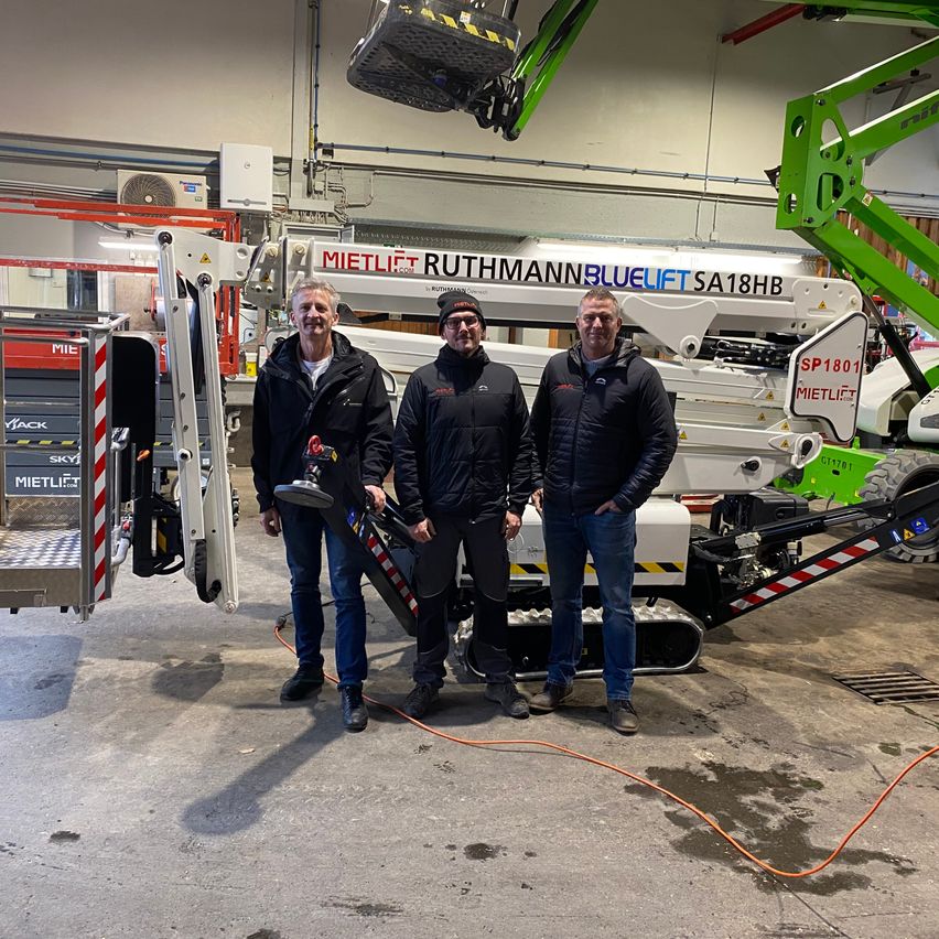 Drei Männer in Wintermänteln stehen vor einem großen, grünen, mobilen Kran in einem Lagerhaus. Ein Mann hält ein Werkzeug in der Hand.