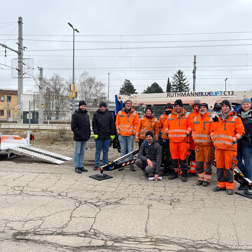 A group of people in orange safety suits pose for a photo with a crane vehicle in the background.