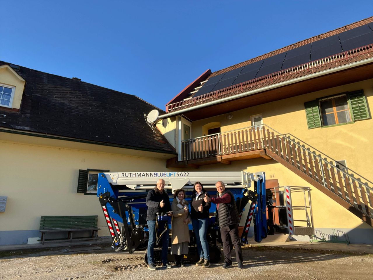 Vier Personen stehen vor einer blau-weißen Maschine und lächeln. Sie geben Daumen hoch. Die Maschine trägt den Text 'RUTHMANNBLUELIFT SA22'. Dahinter befindet sich ein Gebäude mit Balkon.