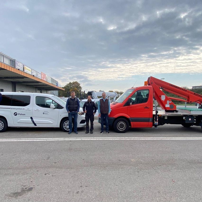 Drei Männer stehen vor einem Parkplatz neben einem weißen Van und einem roten Lastwagen mit ausgefahrenem Kran. Im Hintergrund befindet sich ein Gebäude mit Schildern und einem bewölkten Himmel.
