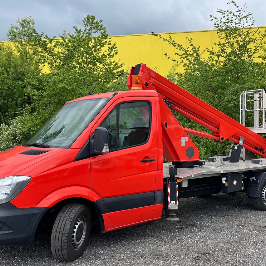 Ein roter Lkw mit einer Scherenarbeitsbühne steht auf einem Kiesplatz mit grünen Bäumen und einem gelben Gebäude im Hintergrund.