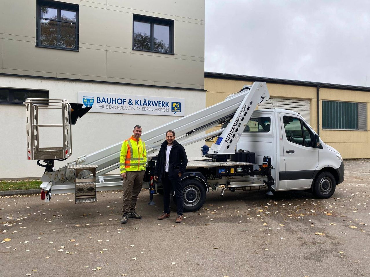 Two men stand in front of a large truck with an extended cherry picker. Behind them is a building with a sign reading 'Bauhof & Klarwerk'.