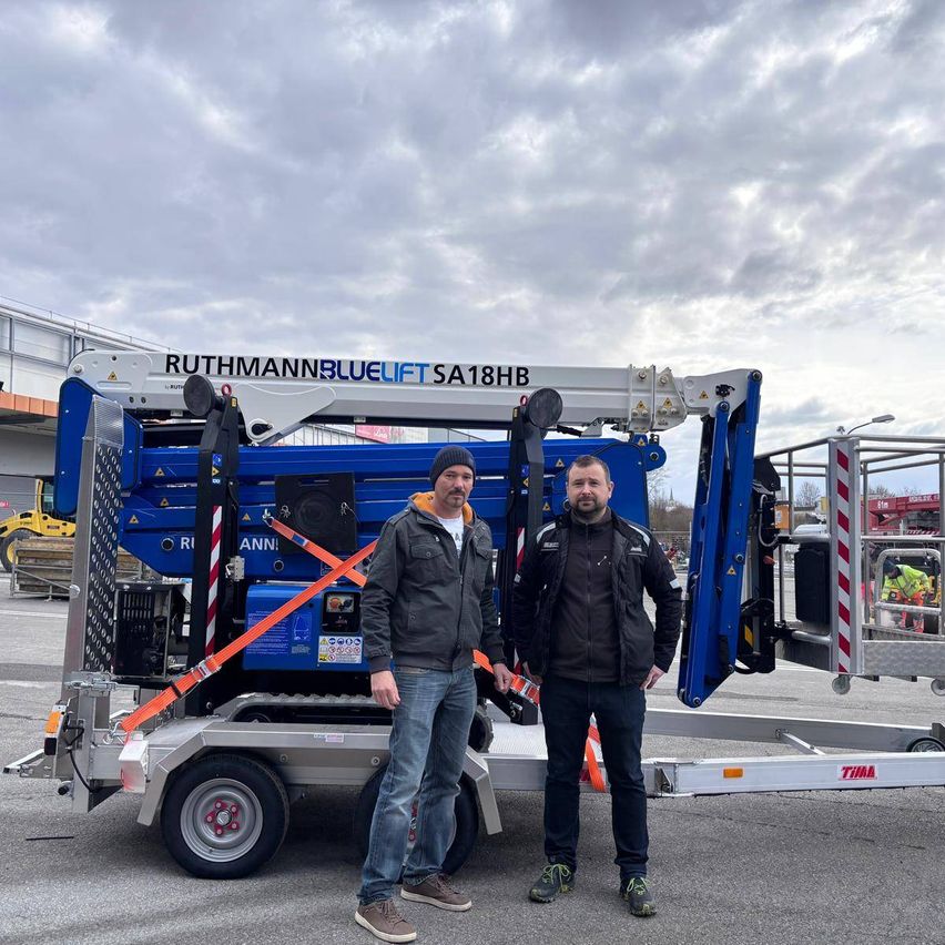 Two men stand in front of a blue and white machine with the words Ruthmann Bluelift SA18HB. The men are wearing jackets and sneakers, and the machine is on a trailer.