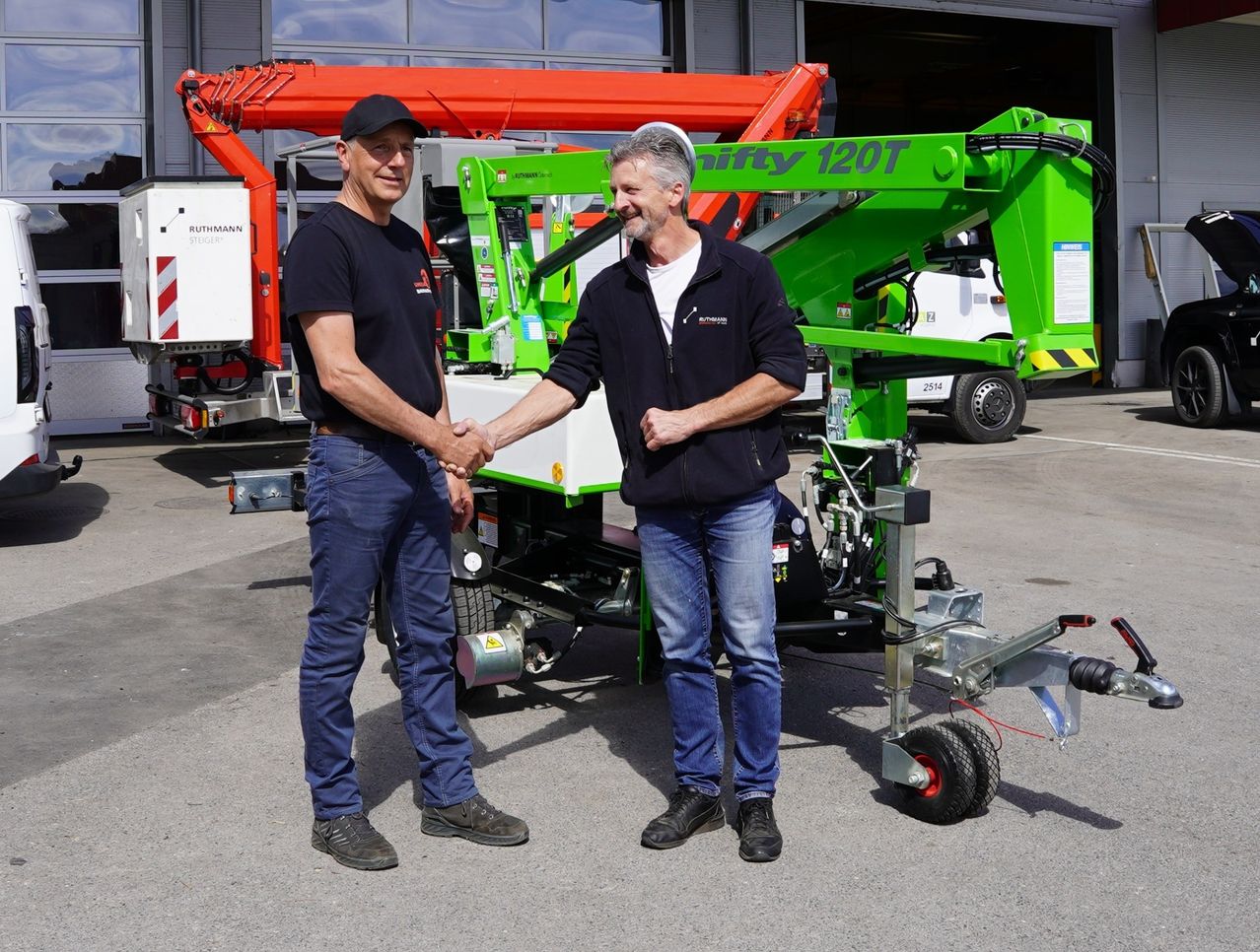 Two men are standing and shaking hands in front of a green machine. Behind them, there is a red and white vehicle.