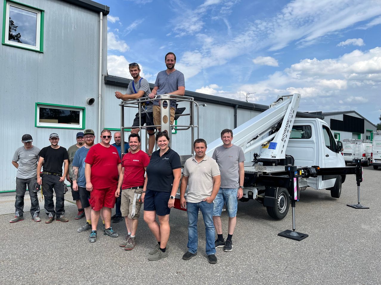 A group of people, including two standing on a cherry picker, pose for a photo in front of a building and a white truck.