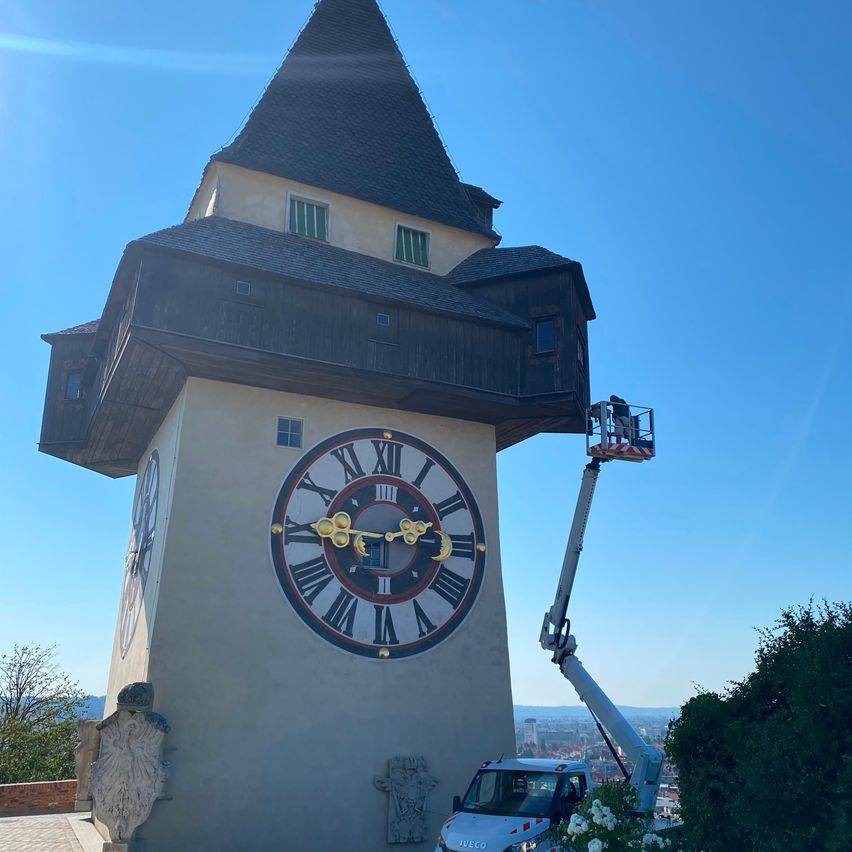 Ein Uhrturm mit einer großen Uhr und einer Person auf einer Plattform, die ihn reinigt. Ein weißer Lastwagen mit einem Kran steht in der Nähe.