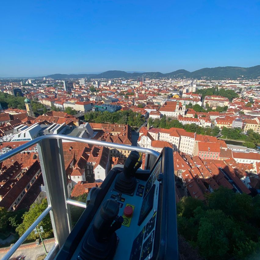 Ein Panoramablick auf eine Stadt mit vielen Gebäuden, einige mit roten Dächern, unter einem klaren blauen Himmel. Im Vordergrund befindet sich ein Kontrollpanel mit Hebeln und Tasten.