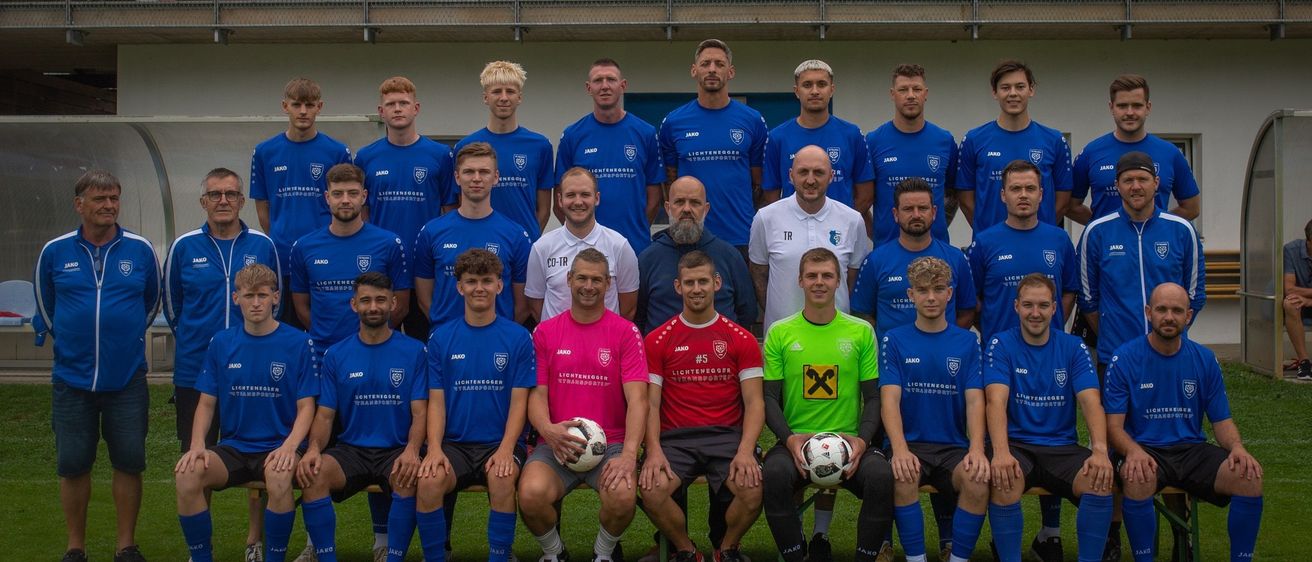 Eine Fußballmannschaft in blauen Trikots posiert für ein Gruppenfoto auf einem grünen Feld, wobei zwei Spieler vorn Fußballbälle halten, und mehrere Personen sitzen auf den Tribünen im Hintergrund.
