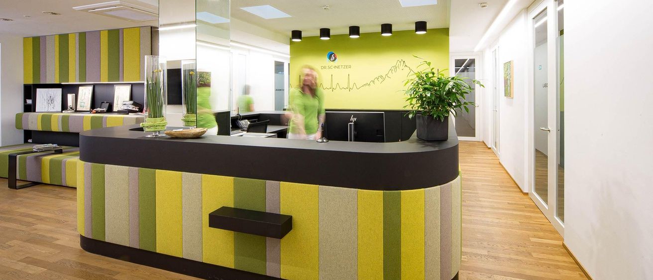 Eine moderne Bürolobby mit einer geschwungenen Rezeptionstheke mit gelben und grünen Streifen. Hinter der Theke ist eine Frau in einem grünen Shirt verschwommen im Hintergrund zu sehen, und auf dem Schreibtisch steht ein Monitor.