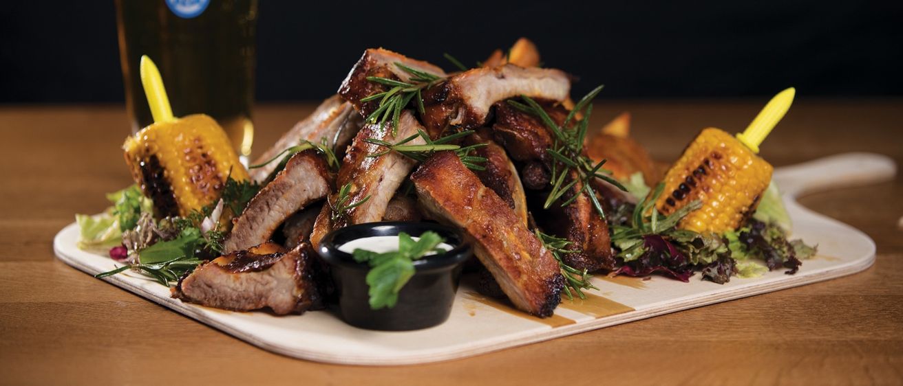 Grilled ribs with rosemary on a white plate, accompanied by corn on the cob and a small dip. A glass of beer with a blue label is also on the table.