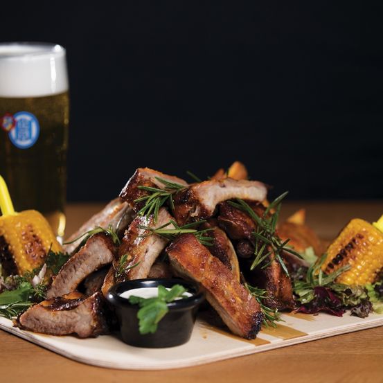A wooden table displays a plate of ribs with herbs, corn, and a salad. Beside it, there is a glass of beer.