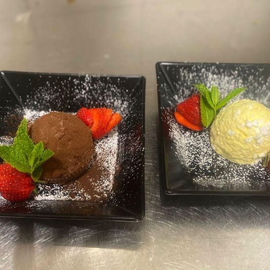 Two black square plates with desserts. One has a chocolate ice cream with chocolate sauce, strawberries, and mint leaves. The other has vanilla ice cream with powdered sugar, strawberries, and mint leaves.