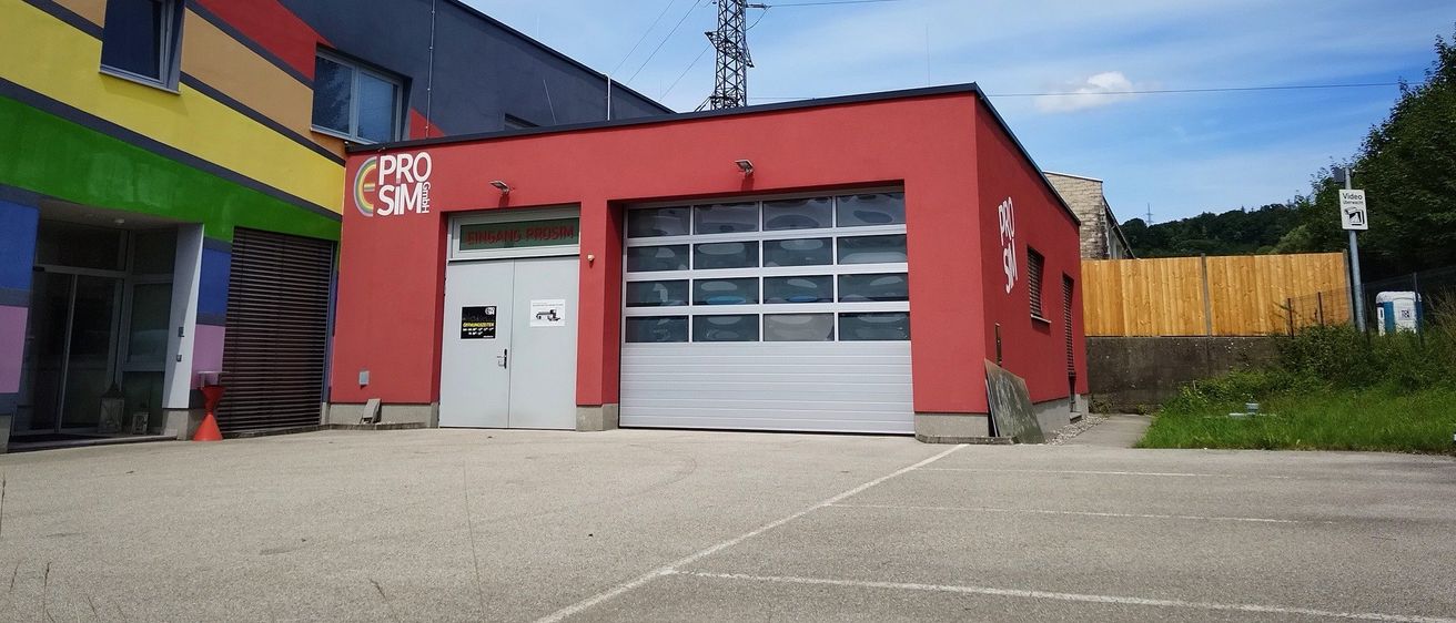 Ein rotes Gebäude mit einer großen Garagentür und dem Logo 'PRO SIM' ist an einem sonnigen Tag zu sehen.