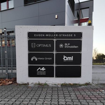 Eine Betonwand zeigt mehrere Firmenschilder vor einem modernen Gebäude. Die Schilder tragen Namen wie 'Optimus', 'BLP Immobilien' und 'Interior GmbH'. Die Wand befindet sich in der Eugen-Müller-Strasse 5.