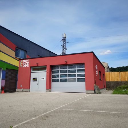 Ein rotes Gebäude mit dem Namen 'Pro Sim' ist sichtbar und verfügt über eine Garage mit großen Fenstern und einem Logo an der Seite. Das Gebäude hat ein Flachdach, und im Hintergrund sind Stromleitungen zu sehen.