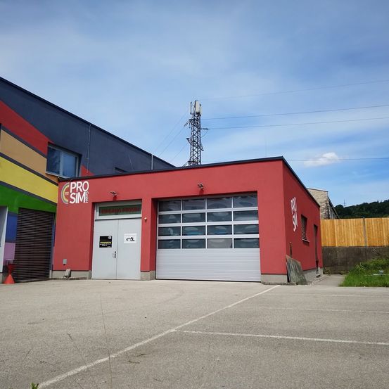 Ein rotes Gebäude mit dem Namen 'Pro Sim' ist sichtbar und verfügt über eine Garage mit großen Fenstern und einem Logo an der Seite. Das Gebäude hat ein Flachdach, und im Hintergrund sind Stromleitungen zu sehen.