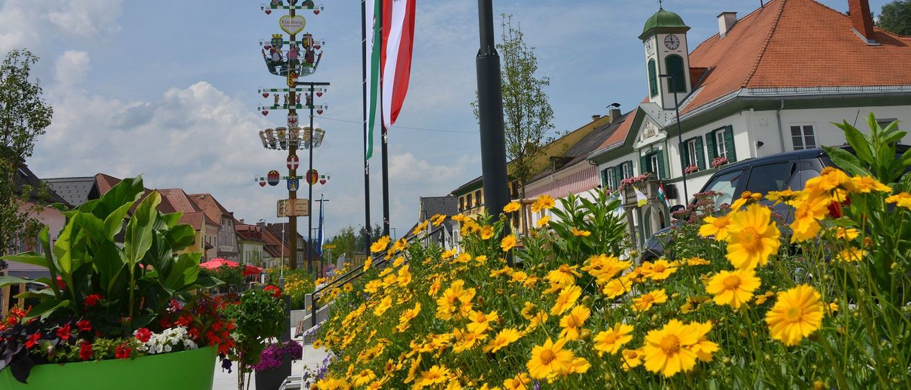 Eine lebendige Straßenszene mit einem Fahnenmast, bunten Blumen und traditionellen Gebäuden. Ein Turm mit einer Uhr steht in der Ferne.