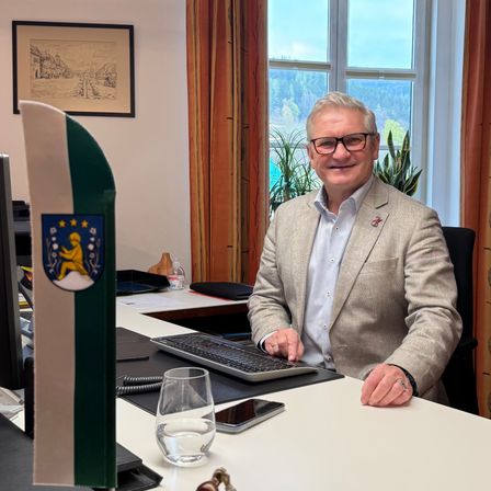 Ein Mann sitzt in einem Büro an einem Schreibtisch und lächelt in die Kamera. Eine Flagge und Trophäen sind sichtbar.