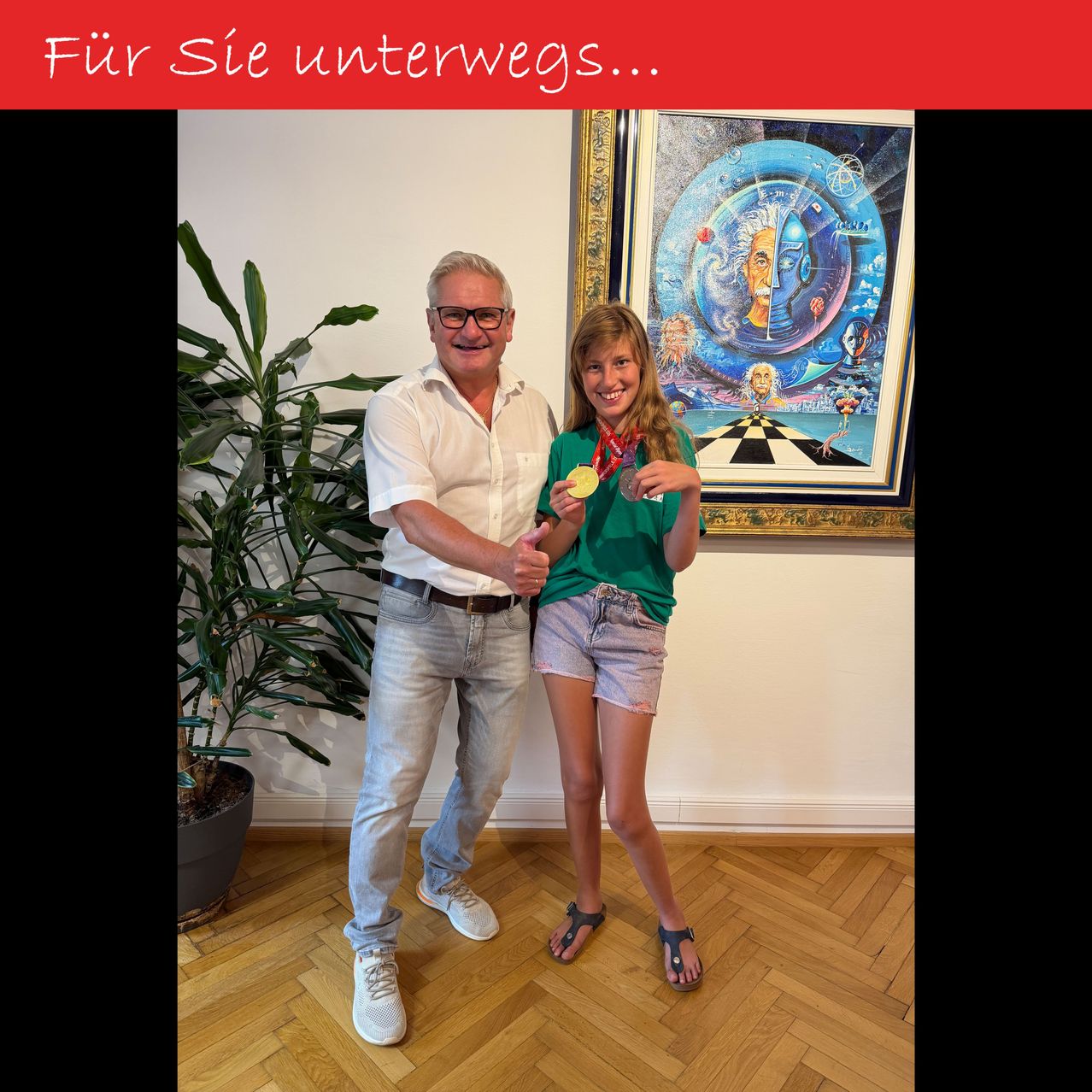 Ein Mann und ein Mädchen stehen lächelnd zusammen in einem Raum und halten goldene Medaillen. Das Mädchen trägt ein grünes Shirt und Shorts, und der Mann trägt ein weißes Hemd, Jeans und eine Brille. Hinter ihnen hängt ein Gemälde an der Wand.