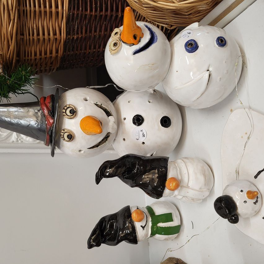 Eine Sammlung von Keramik-Schneemännern, die auf einer weißen Oberfläche ausgestellt sind, darunter einer mit Hut, Brille und Schal, ein anderer mit großer Nase und andere mit verschiedenen Gesichtsausdrücken.