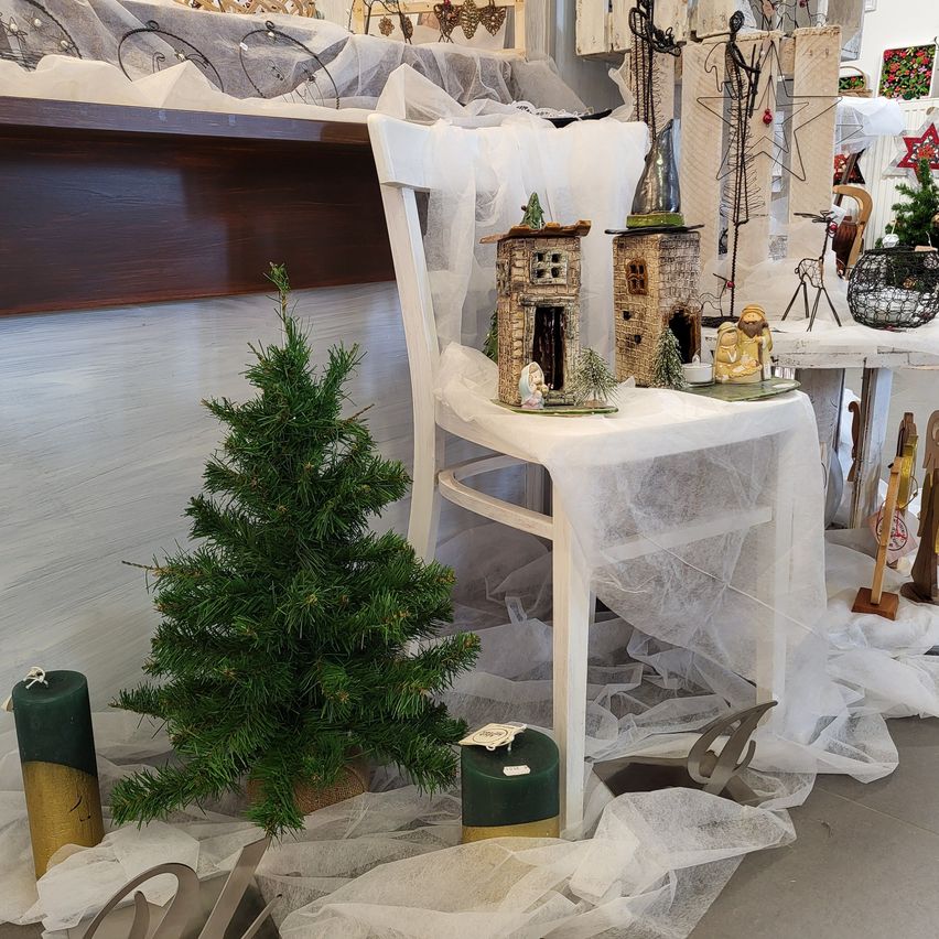 Ein Weihnachtsbaum steht vor einem mit weißem Stoff bedeckten Stuhl. Dahinter befinden sich zwei Miniaturhäuser mit einer Weihnachtsszene und weitere Feiertagsdekorationen.