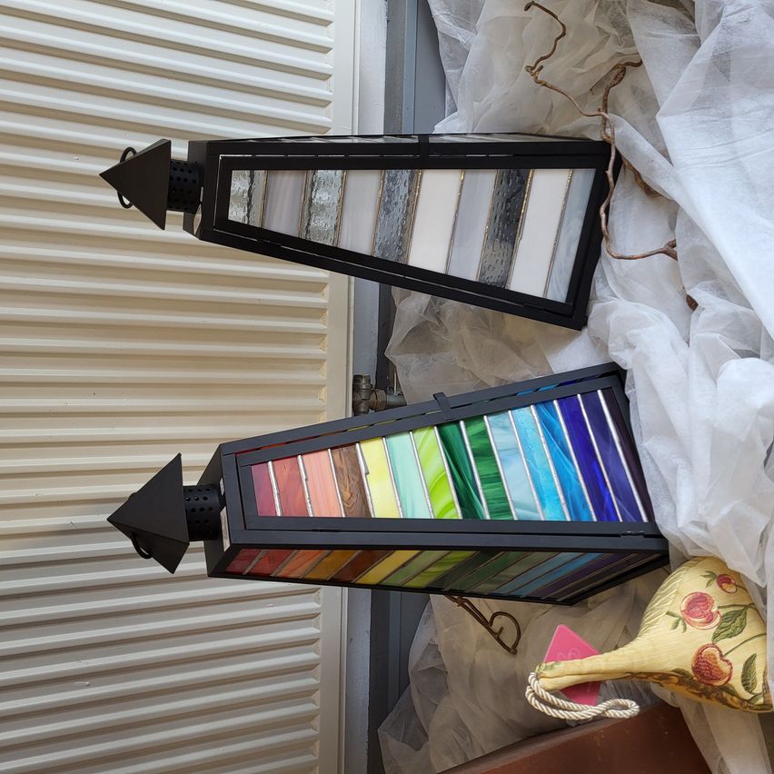 Zwei farbige Glaslaternen mit schwarzen Rahmen liegen auf einem weißen Tuch. Eine Laterne hat ein einzelnes Pfeildesign, während die andere ein mehrfarbiges Design hat. Rechts ist ein gelbes Blumenkissen.