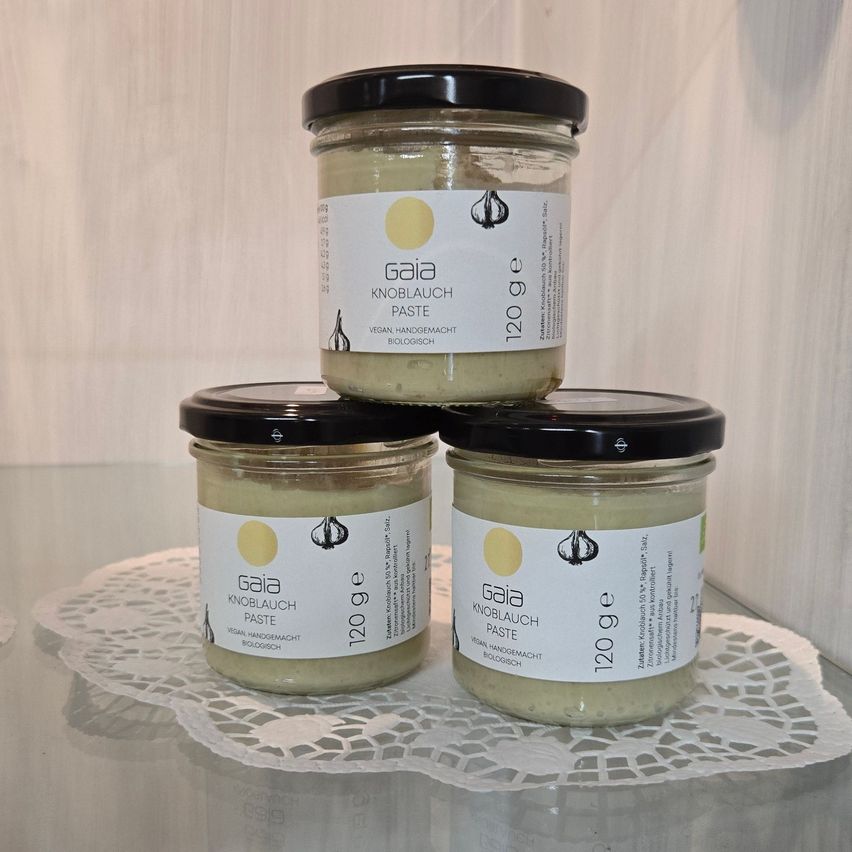 Drei Gläser von Gaia Knoblauch Paste, einer veganen Knoblauchpaste, sind auf einem weißen Doily auf einer Glasoberfläche gestapelt. Jedes Glas hat einen schwarzen Deckel und ein weißes Etikett mit einem Knoblauchsymbol und Text.