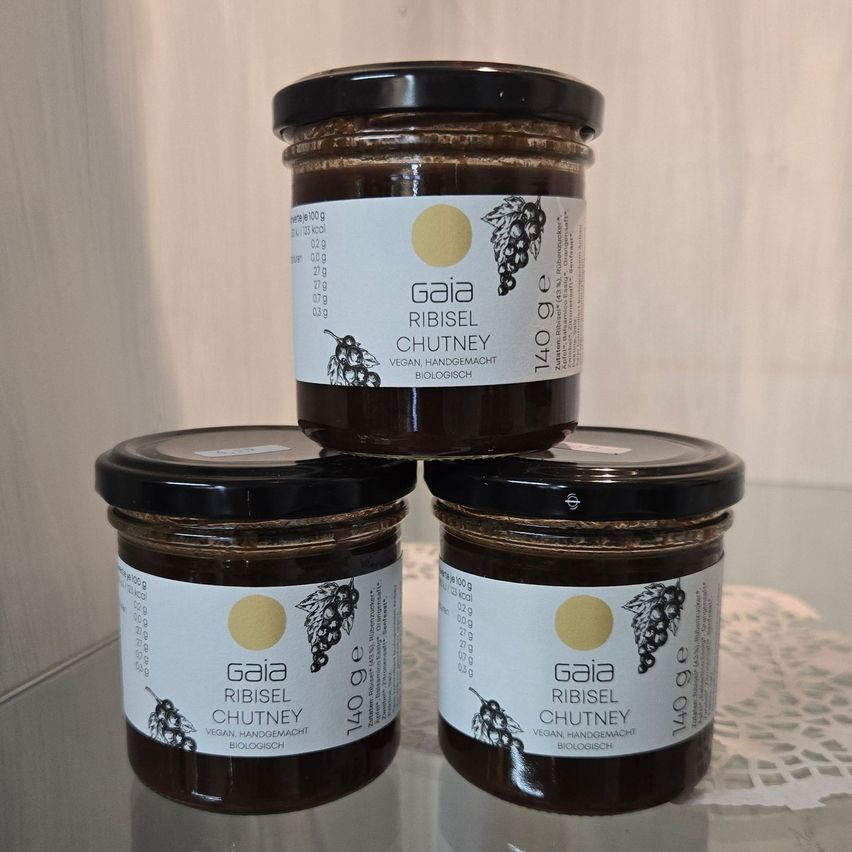 Drei Gläser von Gaia Ribisel Chutney, gestapelt auf einer Glasoberfläche. Jedes Glas hat einen schwarzen Deckel und ein weißes Etikett mit einem gelben Kreis und schwarzem Text.